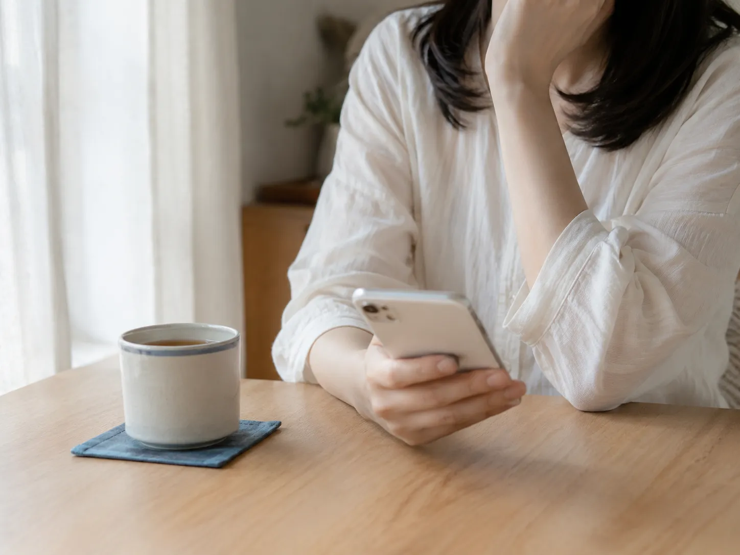 窓辺のテーブルでスマートフォンを見ている女性の手元。湯のみが置かれた明るい部屋